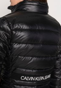 Černá péřová bunda má lesklou, hladkou texturu, horizontální prošití a elastický pásek se značkou v pase s nápisem "CALVIN KLEIN JEANS."