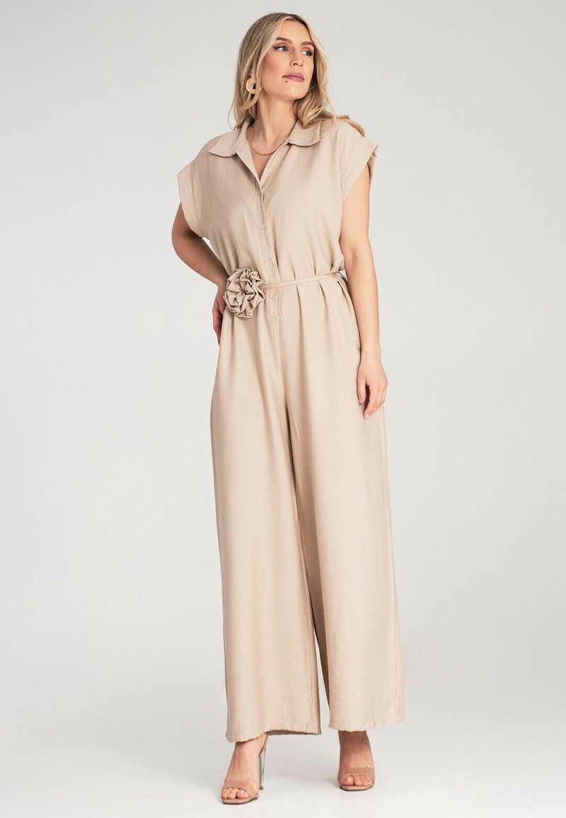 Beżowy kombinezon z kołnierzykiem, krótkimi rękawami, zapięciem na guziki i materiałowym kwiatem akcentującym w talii. Luźny, szeroki krój nogawek.