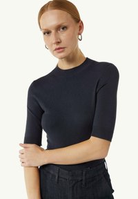 Granatowy dopasowany sweter z krótkimi rękawami, z prążkowanym dekoltem i dołem. Model nosi ciemne spodnie, eksponując gładką teksturę swetra i prosty design.