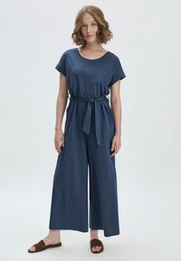 Combinaison bleu marine à manches courtes, avec une ceinture à la taille, un design à jambes larges et une texture de textile lisse. Associée à des sandales marron.