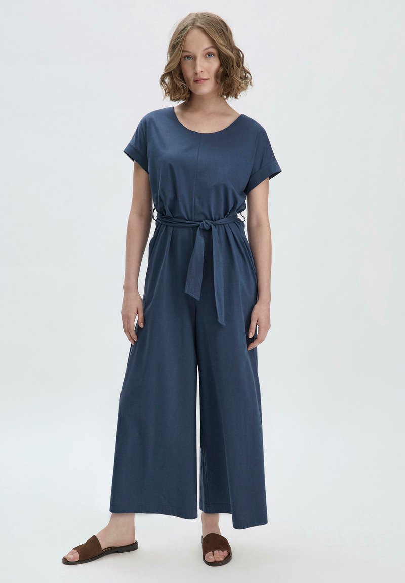 Combinaison bleu marine à manches courtes, avec une ceinture à la taille, un design à jambes larges et une texture de textile lisse. Associée à des sandales marron.
