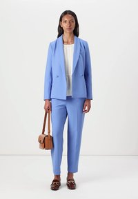 Lyseblå blazer og matchende skreddersydde bukser laget av glatt stoff, kombinert med en plisserte hvit topp og en brun skulderveske.