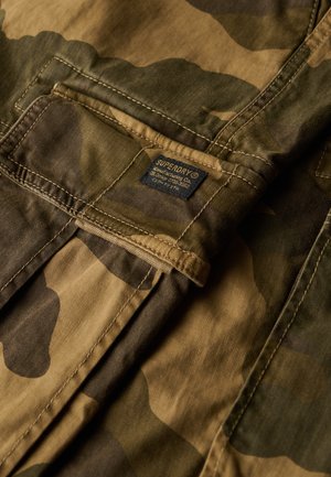 Camo-patroon stof in aardetinten met een zachte textuur. Heeft een label met "Superdry" in het blauw. Het ontwerp omvat zichtbare stiksels en zakken.