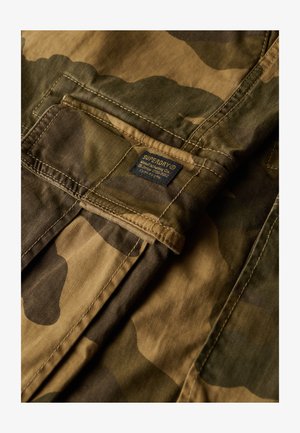 Camouflage-mønstret stof i jordfarver med en blød tekstur. Har et mærke med "Superdry" i blå. Designet inkluderer synlige sømme og lommer.