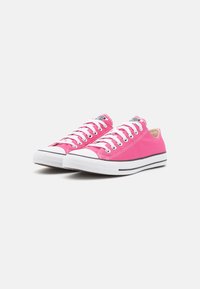 Pink canvas sneakers with white rubber soles and toe caps, featuring white laces, black accents, and a logo label on the tongue.