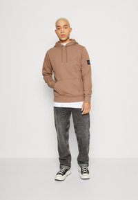 Felpa marrone con cappuccio e lacci, tasca frontale e patch del logo sulla manica, indossata sopra una camicia bianca; abbinata a jeans grigi e sneakers.