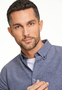 Blauwe chambray knoopshirt met een kraag, een borstzak en contrasterende knopen, gedragen over een wit T-shirt. Close-upportret van een man.