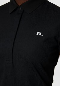 Czarna koszulka polo z teksturowaną powierzchnią, z zapięciem na guziki przy kołnierzyku oraz białym logo po lewej stronie klatki piersiowej. Materiał wydaje się gładki i lekki.