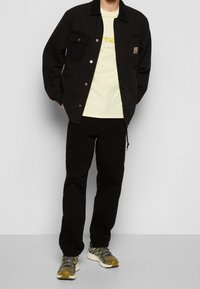Svart jacka med två framfickor, ljusgul t-shirt med logotyp, svarta byxor med blixtlås och gråa och gula sneakers.
