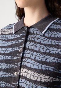 Strikket cardigan med en grå krave, der har et blå og cremefarvet bladmønster på en mørk baggrund, fremhævet af metalliske accenter.