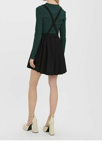 Zwarte plooienrok met een aansluitend groen langemouwtop, voorzien van een gekreukeld textuur en zwarte schouderbanden die op de rug gekruist zijn. Beige blokhak schoenen.