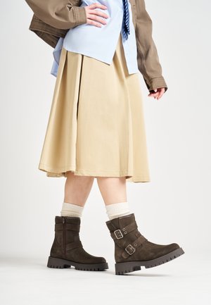 Botas de ante marrón hasta el tobillo con dos hebillas, suelas de goma antideslizantes, combinadas con una falda beige y calcetines de punto crema.