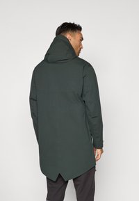 Mørkegrøn lang jakke med hætte, der har en lige snit, glat tekstur og design med høj-lav kant. Inkluderer minimalistische detaljer.