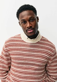 Red and cream patterned sweater with a ribbed collar, featuring a textured knit design and a relaxed fit, worn over a light-colored shirt.