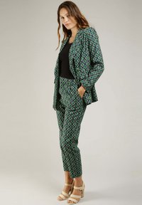 Ensemble de blazer et pantalon à motif vert et bleu marine, avec un design géométrique, en tissu lisse et avec des poches, associé à des talons de couleur claire.