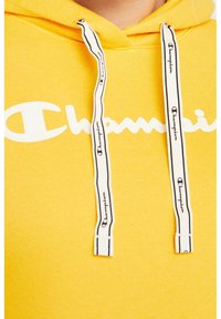 Sweat à capuche jaune avec un grand logo "Champion" blanc. Équipé de cordons blancs avec texte noir. Tissu doux et texturé.