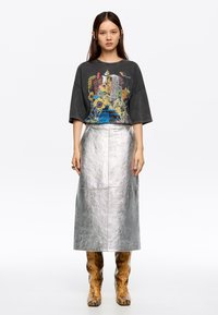 Grafische grijze oversized t-shirt met kleurrijke stads- en bloemenprint, gecombineerd met een metalen zilveren rok tot de knie en gebloemde bruine laarzen.