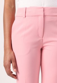 Pantalons tailleurs roses avec une texture lisse, dotés d'une ceinture et de poches latérales, capturés de la taille, main positionnée à proximité.