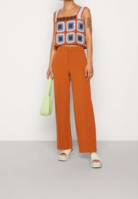 Virkat crop top i orange och blå rutor, parat med vida orange byxor. Håller en strukturerad grön väska, bär beige plattformsandaler.