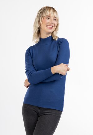 Młoda kobieta o blond włosach uśmiechająca się, ubrana w niebieski sweter z golfem i długim rękawem oraz ciemne spodnie, z założonymi na piersiach rękami.