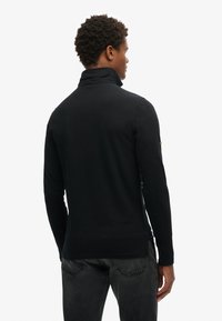 Camisa polo negra de manga larga con cuello alto, diseño ajustado y lados sin costuras, con tela texturizada y detalles minimalistas.