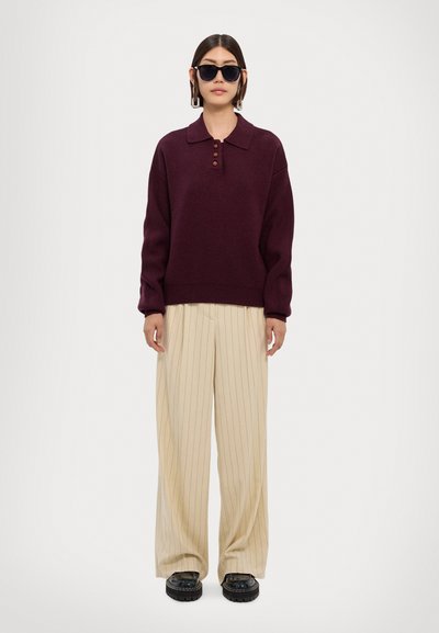 Suéter de punto burdeos con cuello y tres botones, combinado con pantalones de pierna ancha beige con rayas y zapatos negros gruesos.