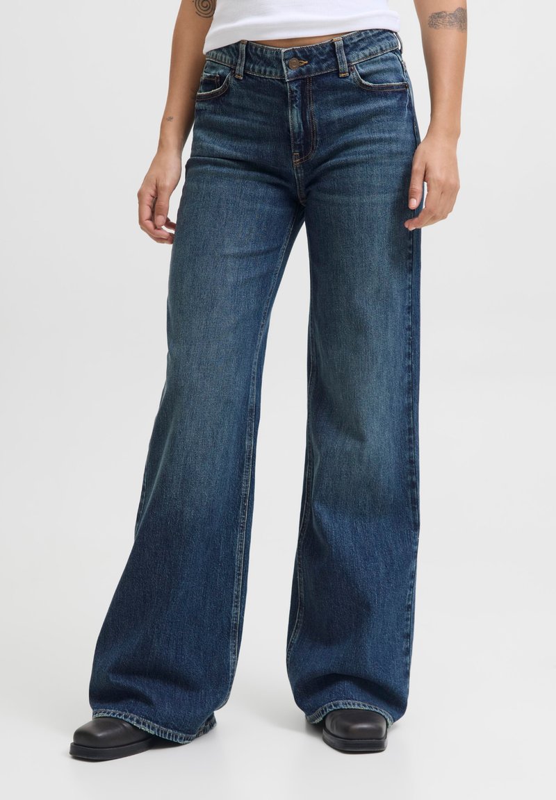 Personne portant un jean large bleu foncé, des chaussures noires et un haut blanc, debout les mains détendues le long du corps.