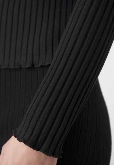 Black ribbed fabric close-up featuring a fitted design with textured surface and flared cuffs at the wrist, showcasing a sleek silhouette.