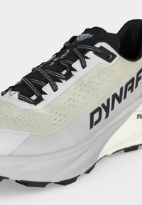 Ανοιχτό γκρι και μαύρο παπούτσι τρεξίματος για μονοπάτια με μαύρα κορδόνια, ανθεκτικό πάνω μέρος από δίχτυ και σήμανση "DYNAFIT" στο πλάι.