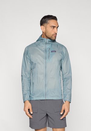 Kerge, veekindel sinine jakk, millel on ees tõmblukk ja kapuuts. Omadused hõlmavad väikest logo rinnal, pikki varrukaid ja siledat tekstuuri.