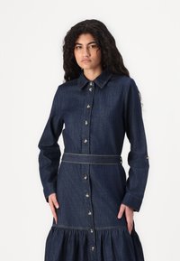 Robe en denim avec un col, des manches longues, des boutons sur le devant, une taille ceinturée et une jupe évasée. Couleur bleu foncé avec des coutures contrastantes.