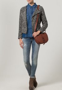 Šedá texturovaná zkrácená bunda s asymetrickým zipem, modrá džínová košile, světlé ošoupané džíny a hnědá crossbody taška. Hnědé kotníkové boty.
