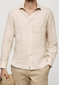 Mężczyzna w beżowej, długorękawowej koszuli zapinanej na guziki z kieszenią na piersi, trzymający w lewej ręce dopasowaną beżową kurtkę, ręce w kieszeniach.