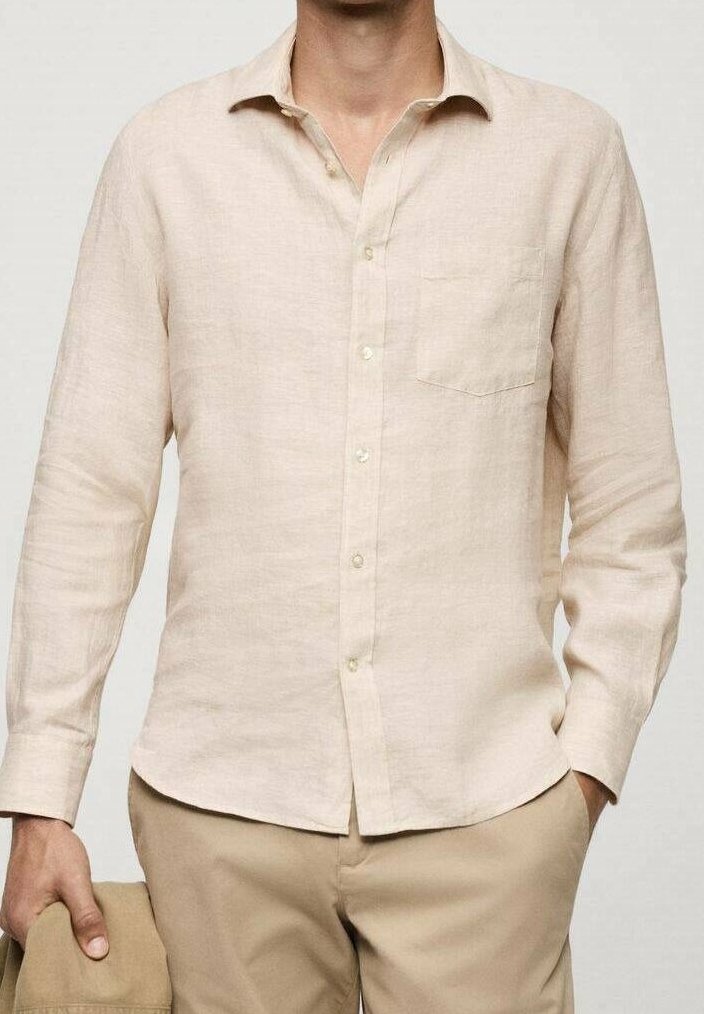 Mężczyzna w beżowej, długorękawowej koszuli zapinanej na guziki z kieszenią na piersi, trzymający w lewej ręce dopasowaną beżową kurtkę, ręce w kieszeniach.