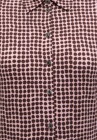 Gros plan d'une chemise boutonnée avec un motif de points irréguliers marron sur un fond rose clair et un col classique.