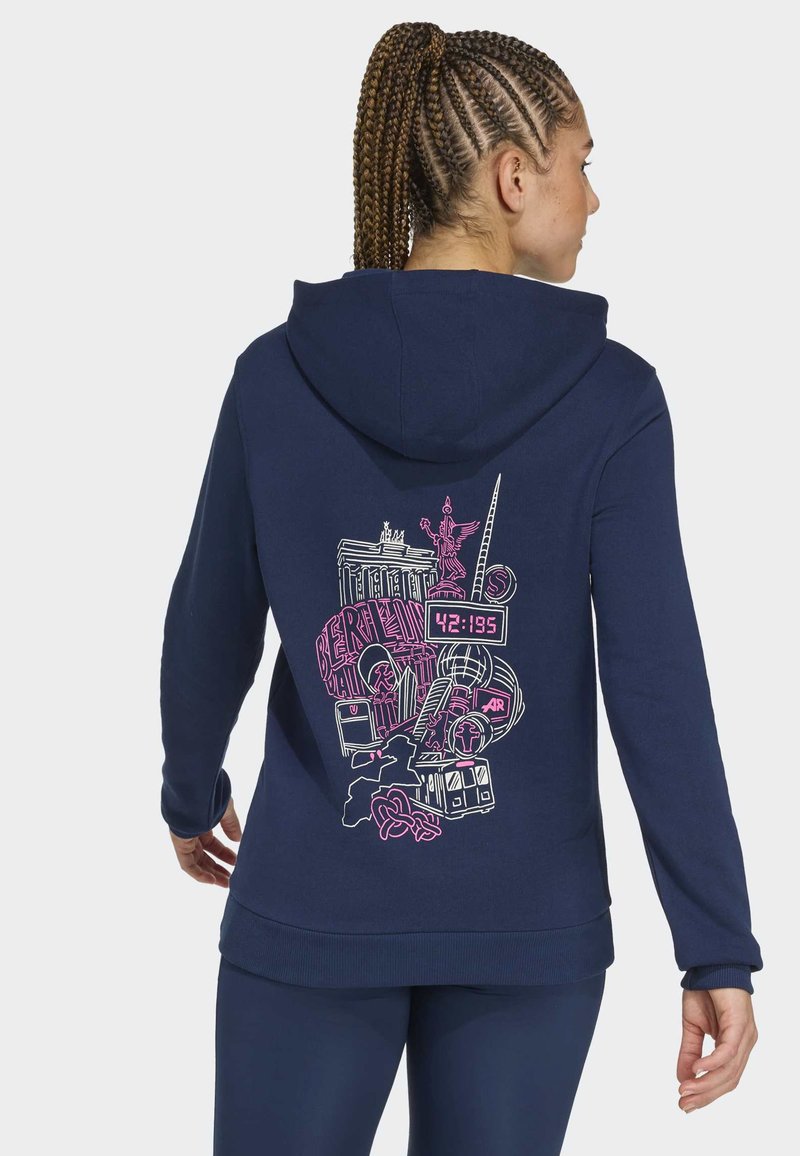 Kvinde med flettet hår iført en marineblå hættetrøje med et pink og hvidt Berlin-tema grafisk design, der inkluderer vartegn og maratonur.