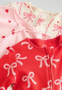 Tre body neonato: uno rosa con cuori rossi, uno crema con fiocchi rossi e uno rosso con fiocchi bianchi. Tessuto in cotone, texture morbida, colletto rotondo.