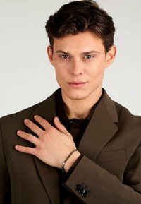 Abito marrone con rever a punta, indossato sopra una camicia scura. Un braccialetto argento e nero è indossato sul polso sinistro, con un design a catena.