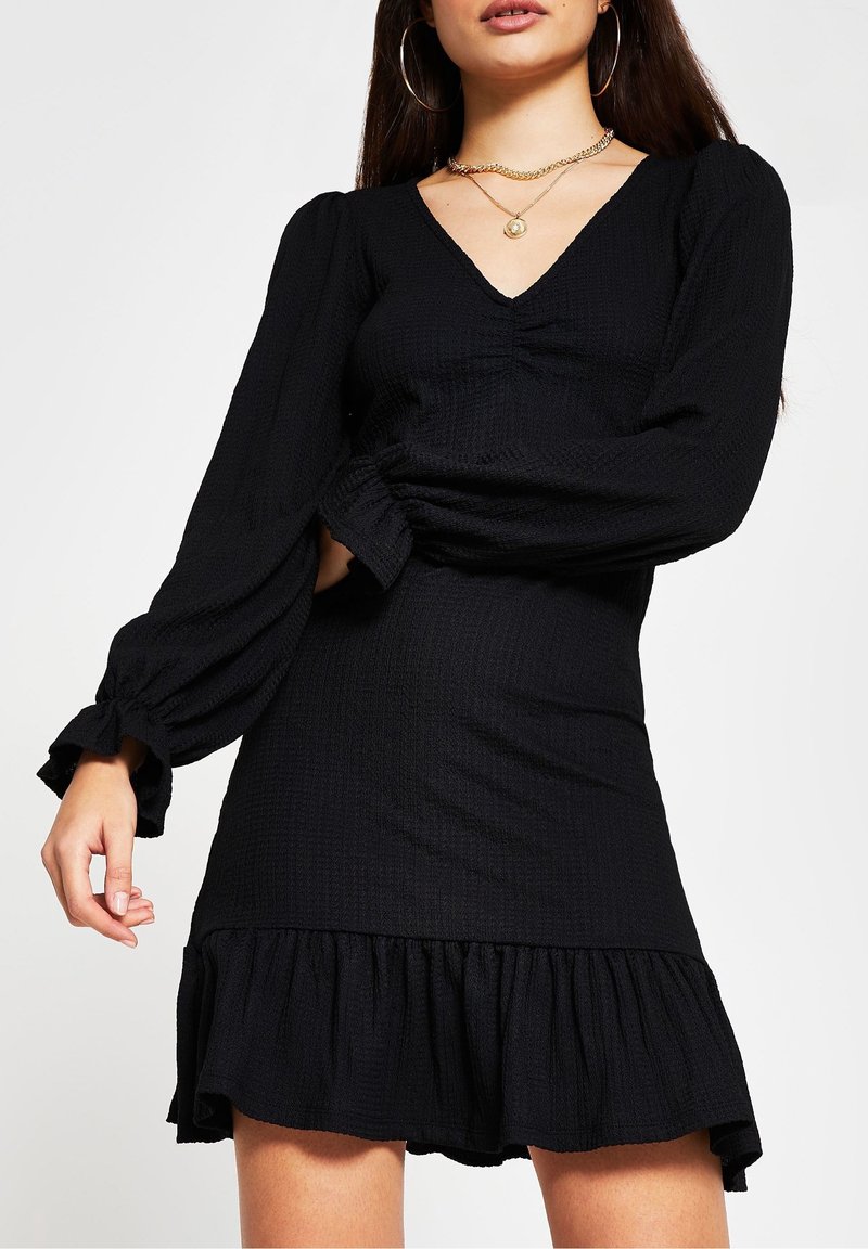 Femme portant une robe noire à manches longues avec un ourlet à volants, un col en V et un tissu texturé, accessoirisée avec des boucles d'oreilles créoles dorées et des colliers superposés.