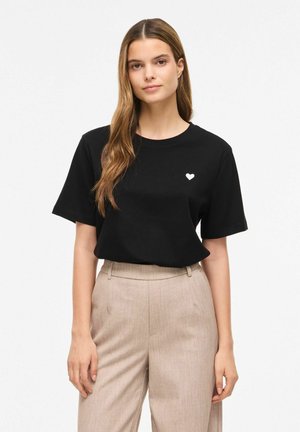 Femme aux longs cheveux portant un t-shirt noir avec un petit cœur blanc sur la poitrine et un pantalon beige taille haute sur un fond uni.