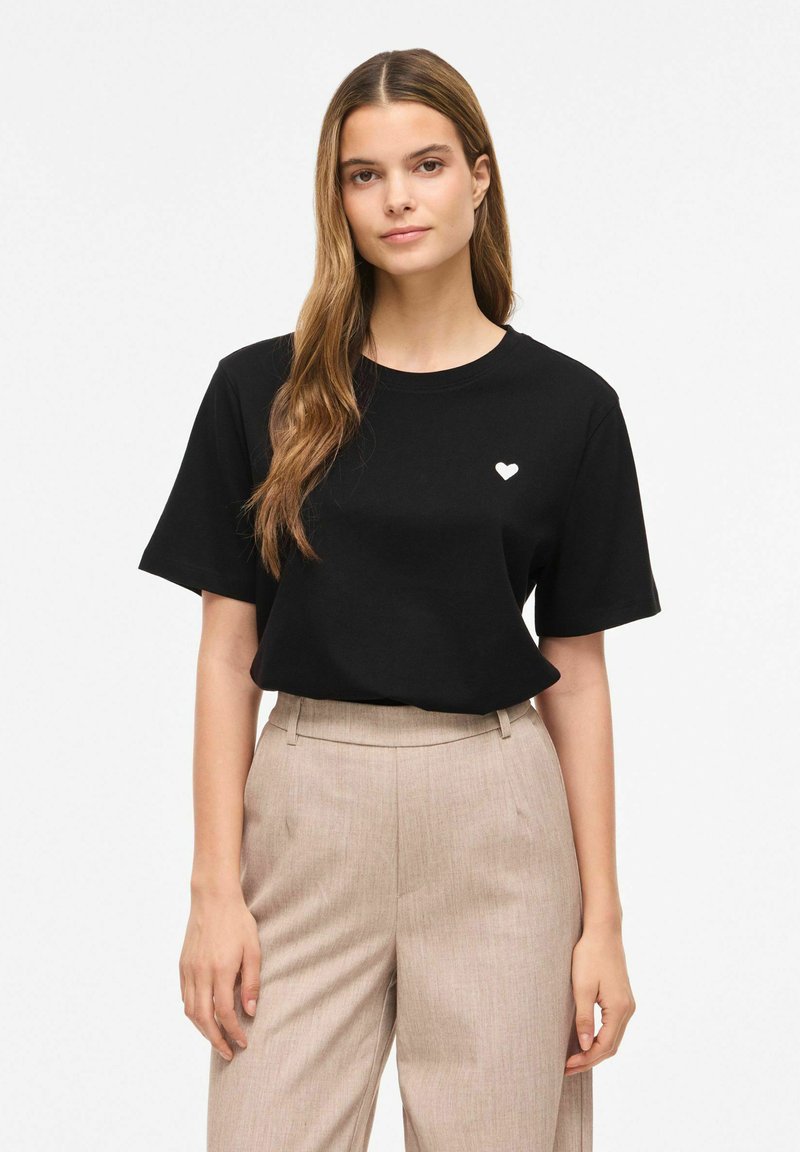 Femme aux longs cheveux portant un t-shirt noir avec un petit cœur blanc sur la poitrine et un pantalon beige taille haute sur un fond uni.