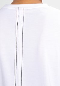 Hvid t-shirt med rund hals, der har to lodrette sorte syninger løbende ned langs midten af ryggen. Blødt, let stof.