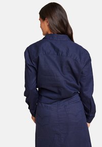 Mørkeblå lin-skjorte med krage og lange ermer, med en tettsittende design og synlige detaljer på baksemen.