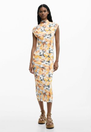 Kvinde iført en ærmeløs midi-kjole med høj hals og et blomsterprint i orange, gult og gråt, stylet med brune plateformesandaler.