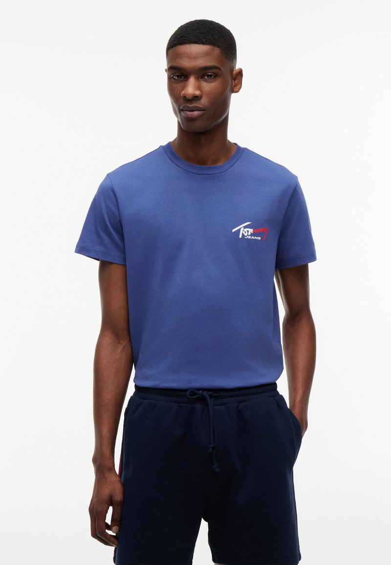 Νέος άνδρας που φοράει μπλε μπλουζάκι Tommy Jeans και σκούρα μπλε σορτς με κορδόνι, στέκεται με το ένα χέρι στην τσέπη μπροστά σε λευκό φόντο.