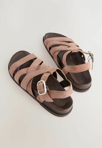 Sandali in suede rosa chiaro con cinturini incrociati, fibbia regolabile alla caviglia e suola piatta marrone scuro. Design testurizzato e minimalista.