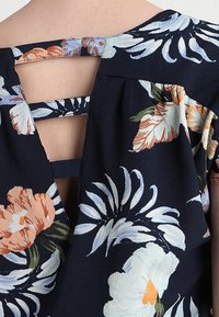 Dos d'un chemisier bleu marine à fleurs orange et blanches avec trois bretelles horizontales en tissu sur un dos ouvert.