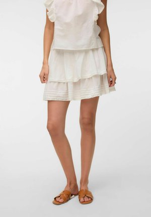 Femme portant une jupe blanche à volants et un haut, assortis à des sandales plates ouvertes couleur beige, debout devant un fond uni.