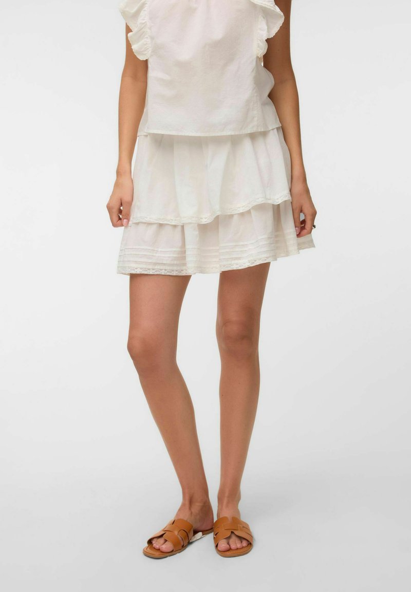 Femme portant une jupe blanche à volants et un haut, assortis à des sandales plates ouvertes couleur beige, debout devant un fond uni.