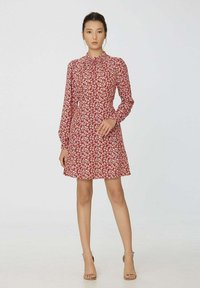 Femme debout dans des talons beiges portant une robe rouge mi-longue avec un motif floral blanc et des manches longues.